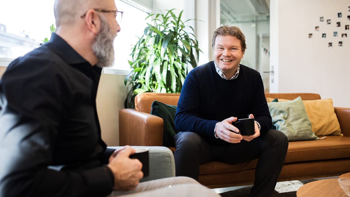 To kollegaer som snakker sammen. De sitter i en sofa og holder i kaffekopper.