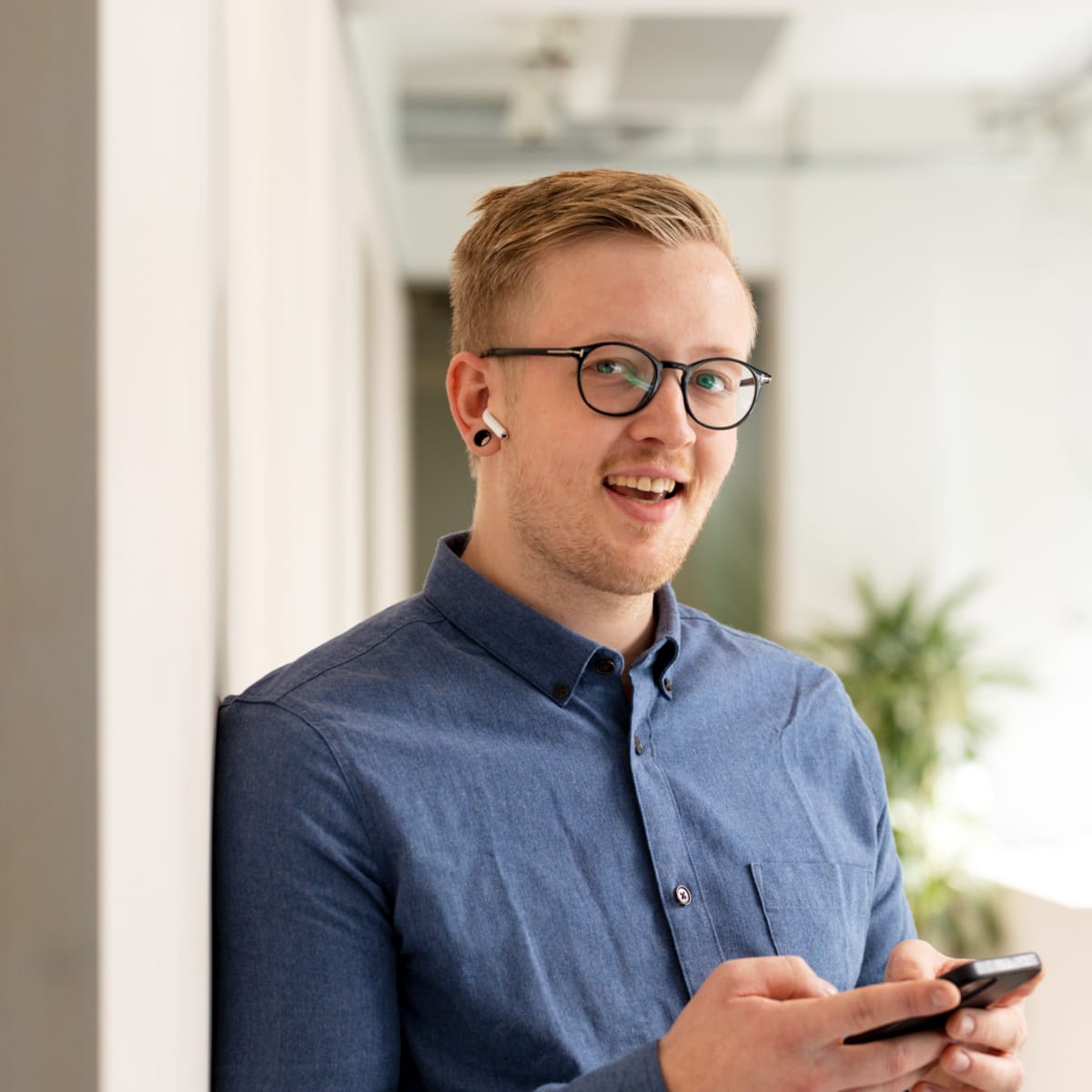 Ansatt som står med mobiltelefon i hånda og airpods i ørene. På seg har han briller med tykk innfatning og blå skjorte.